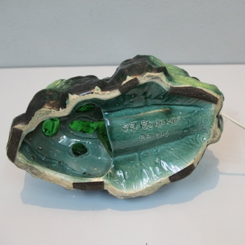 Realizada por el escultor español Santiago Rodrigues Bonome (1901-1995). Sus esculturas están caracterizadas por un gran realismo.
Cristal prensado en verde  y cerámica esmaltada.
Firmada